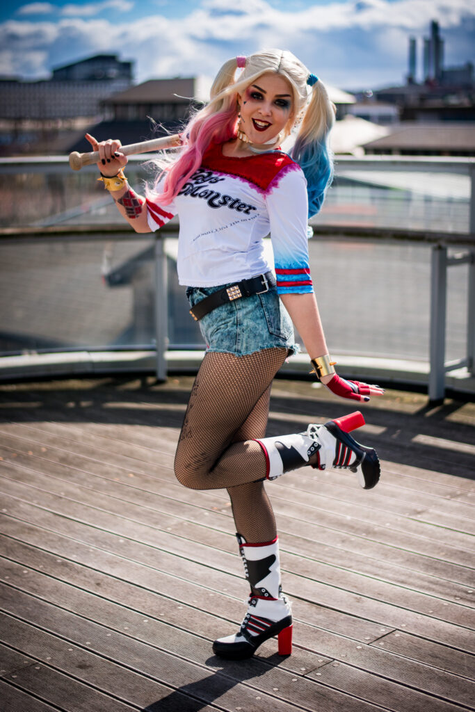 Kami Renee dressed in a detailed Harley Quinn cosplay with colorful pigtails, fishnet stockings, and a baseball bat, posing confidently on an urban bridge under a clear blue sky.