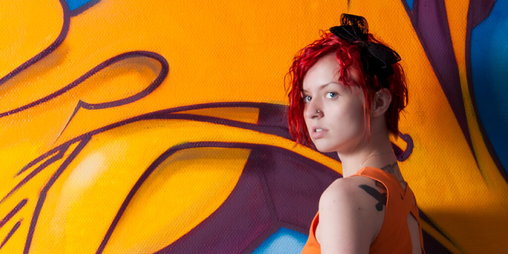 Kybele in an orange dress posing confidently in front of a colorful graffiti background.