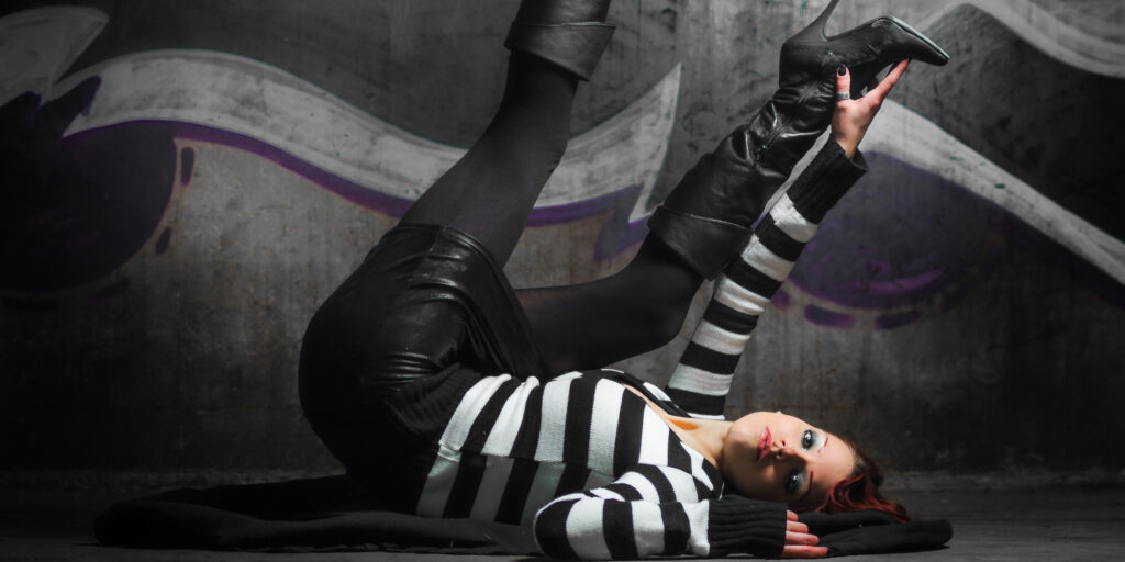 Kiki in an urban setting with graffiti background, wearing a black-and-white striped sweater, leather pants, and black boots in a dynamic pose.