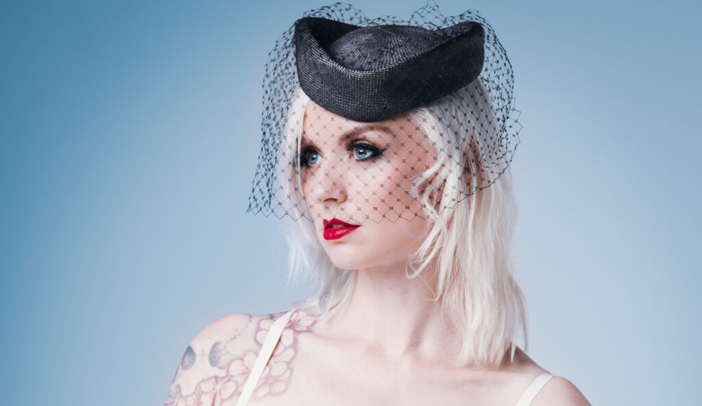 Kybele iført en cremefarvet latexkjole designet af Nicoline Hansen, kombineret med en sort fascinator og hendes tatoveringer, mod en dæmpet blå baggrund.