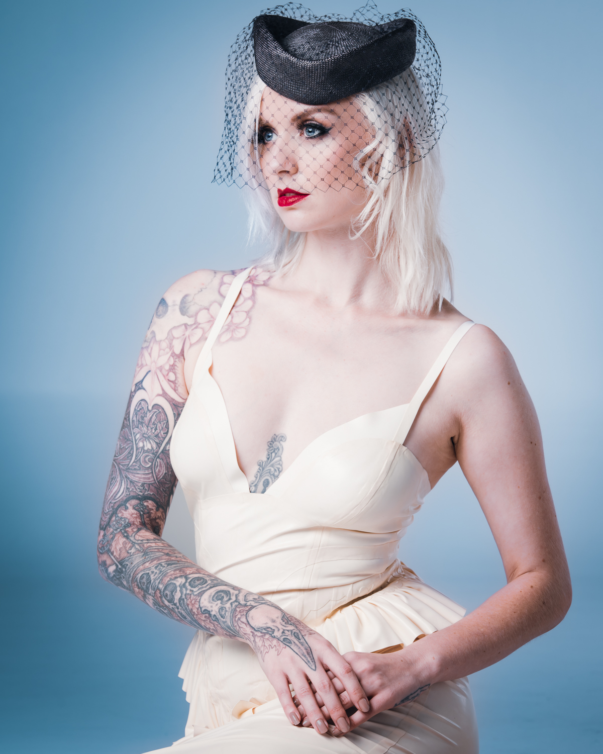 Kybele in a cream latex dress by Nicoline Hansen, paired with a black fascinator and showcasing her tattoos, against a muted blue background.